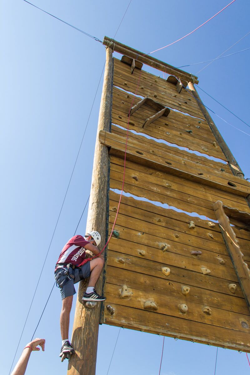 Here is a picture of a person climbing a wooden structure. The person is wearing a helmet and is secured by ropes. The structure appears to be a climbing wall or a similar obstacle. The person is in the process of ascending the wall. The background is a clear blue sky.